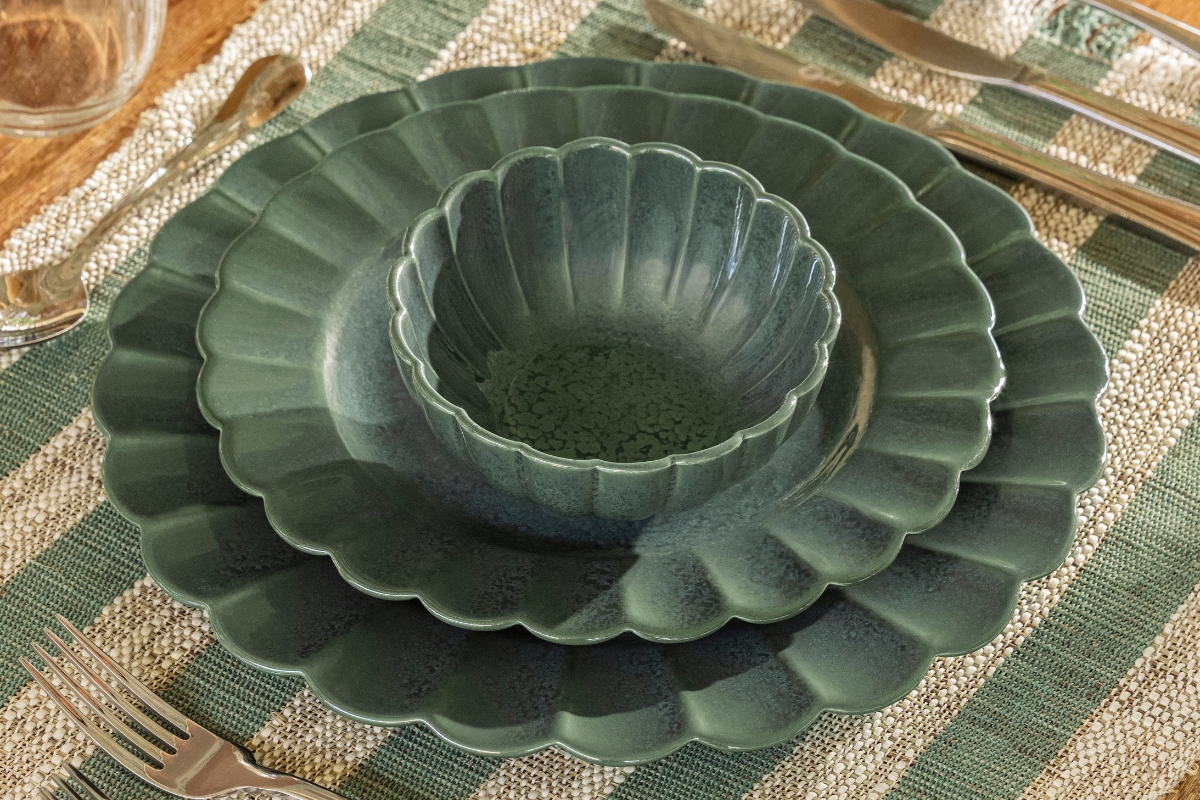 Imagem: Conjunto de louças Oxford em tom verde hortênsia com acabamento brilhoso e bordas em formato de pétalas, disposto sobre jogo americano listrado em tons de verde e bege.
