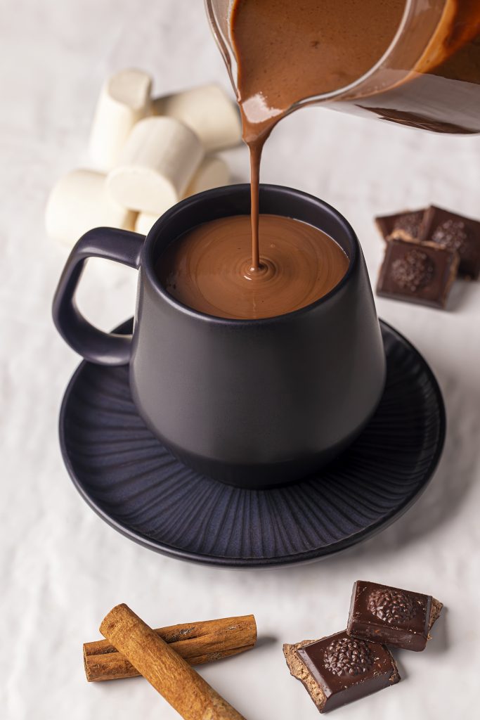 Imagem: Chocolate quente cremoso sendo servido em uma caneca preta com pires combinando.