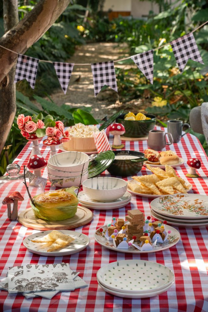 Imagem: ideia de como montar uma mesa de festa junina ao ar livre decorada com toalha xadrez vermelha, louças delicadas, panelas e comidas típicas como pipoca, pastel e bolo, em um ambiente com decoração rústica e cogumelos decorativos.