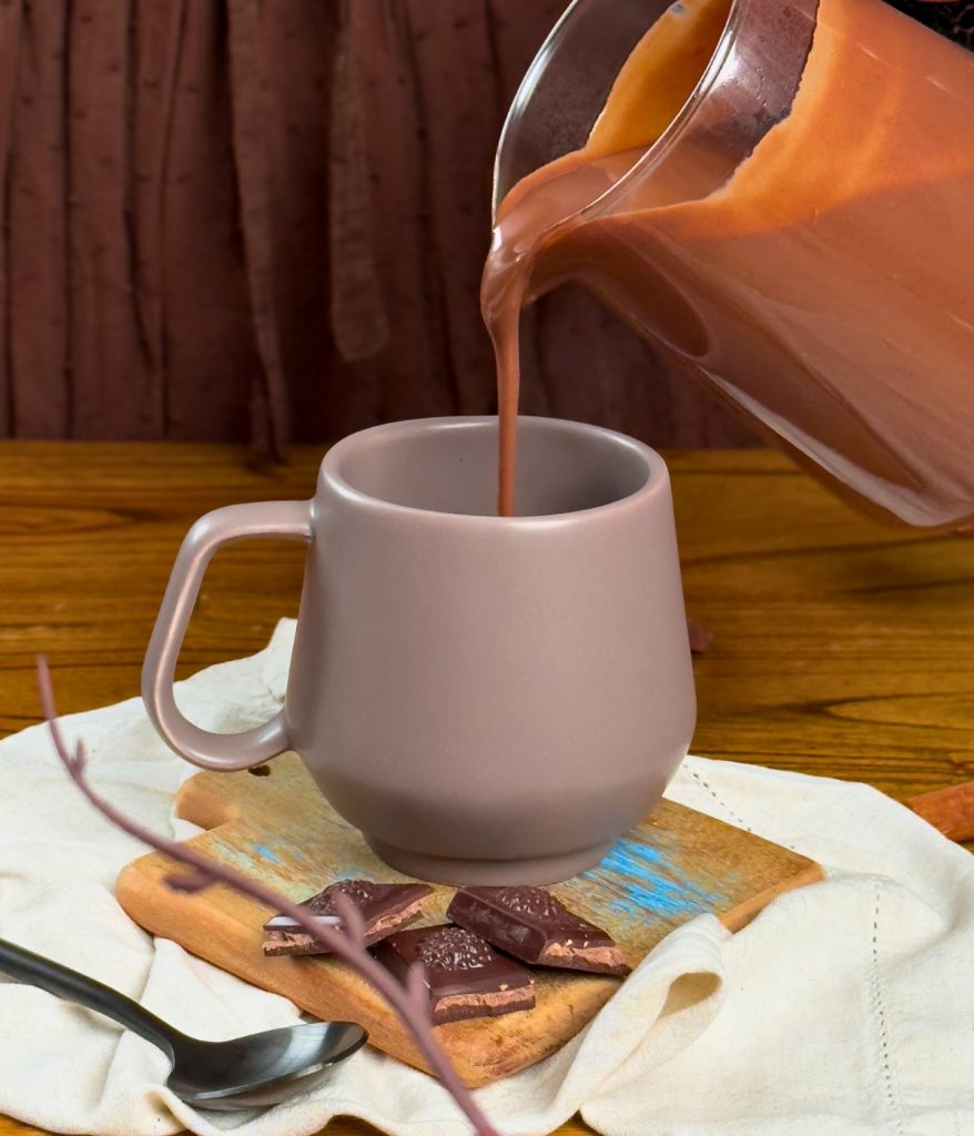 Imagem: Caneca de cerâmica em tom neutro sendo preenchida com chocolate quente cremoso, sobre guardanapo com pedaços de chocolate.
