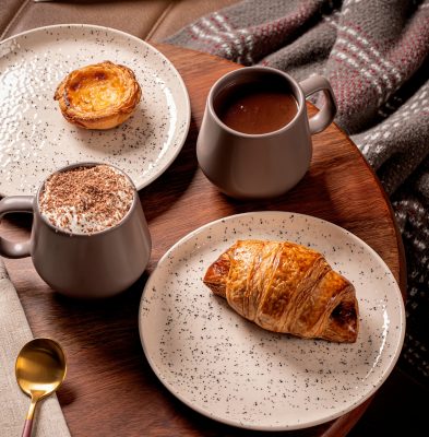 Imagem: Composição de café da manhã com caneca de cerâmica, croissant, pastel de nata e chocolate quente, servidos em pratos com acabamento rústico, sobre mesa de madeira, ao lado de manta xadrez e livro aberto.