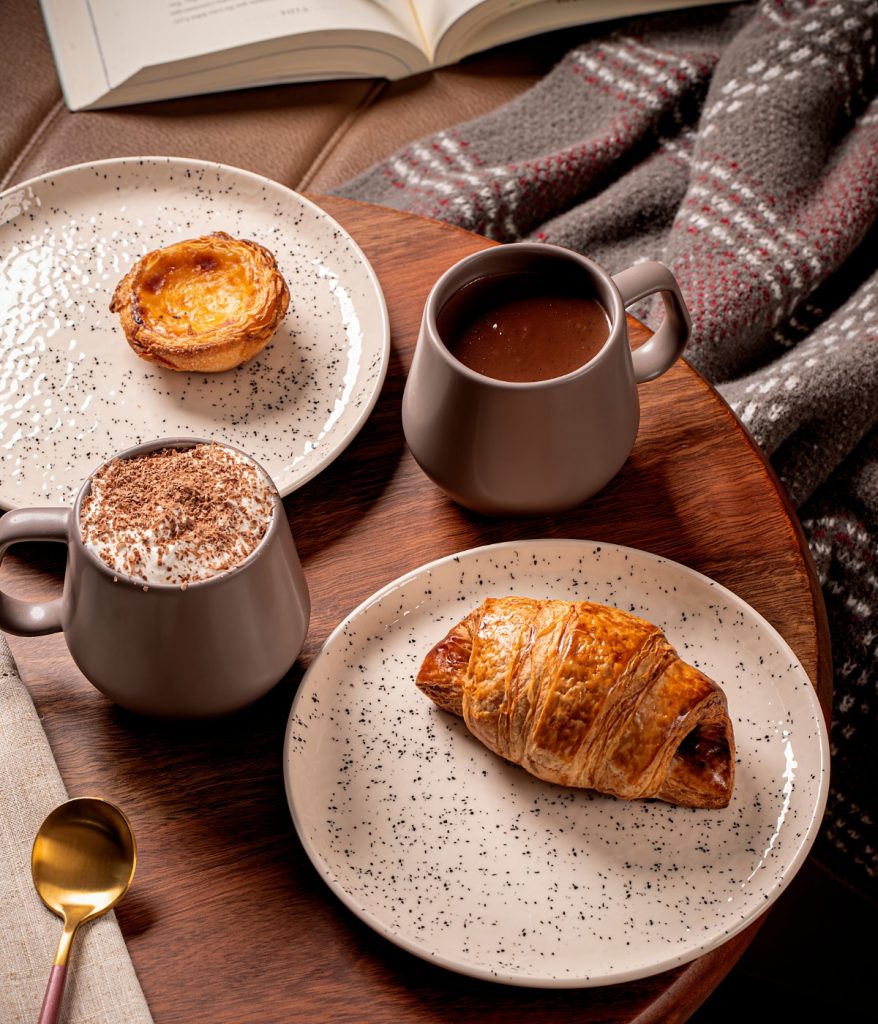 Imagem: Composição de café da manhã com caneca de cerâmica, croissant, pastel de nata e chocolate quente com espuma.
