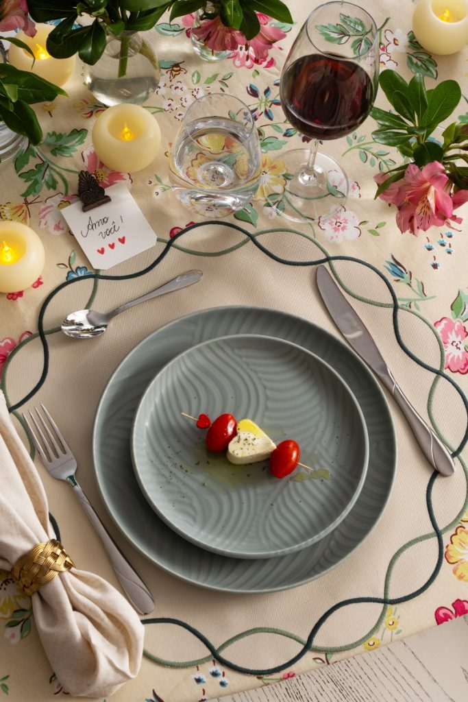 Imagem: Mesa de Dia dos Namorados decorada com toalha floral, pratos verdes com entrada em formato de coração feita de muçarela de búfala e tomates, taça de vinho tinto, copo de água, flores cor-de-rosa, velas acesas e um bilhete escrito “Amo você!” com três corações.