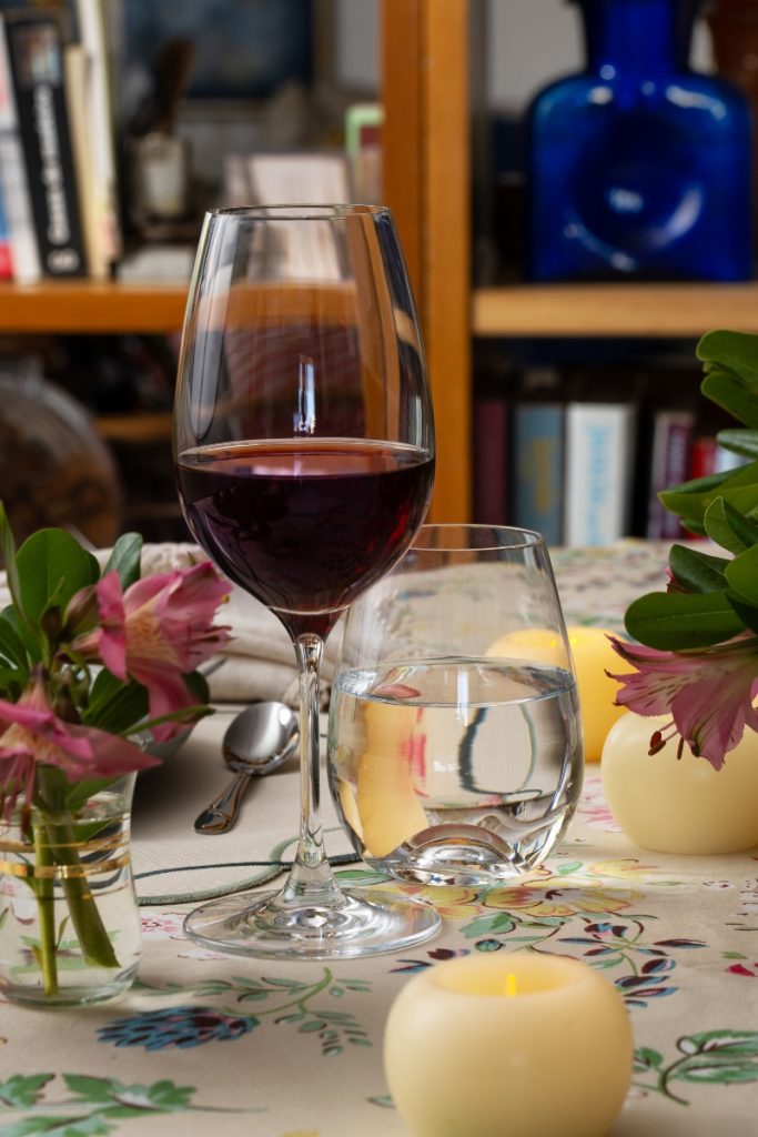 Imagem: Taça de vinho tinto ao lado de copo com água sobre uma toalha com estampa floral. Velas amarelas pequenas e flores cor-de-rosa decoram a mesa, criando um clima íntimo e romântico.