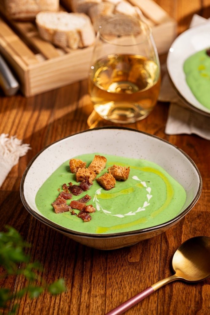 Imagem: Creme de ervilhas com batats servido em bowl da linha Ryo Maresia com croutons, sobre mesa posta com copo de vinho branco e pão.