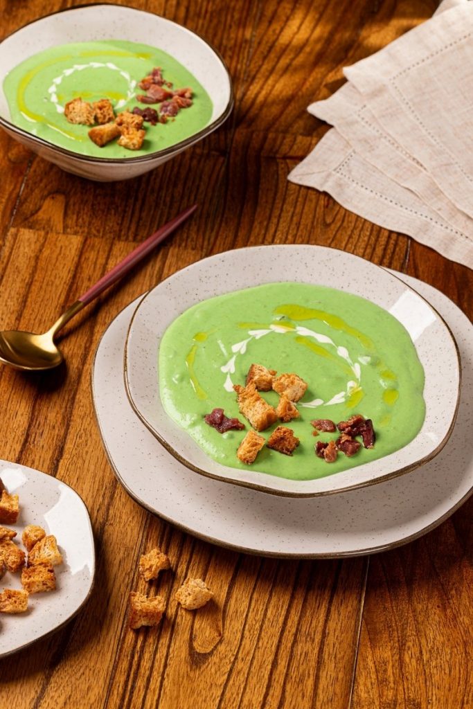 Imagem: Creme de ervilhas com batatas servido em conjunto de tigela e prato da linha Ryo Maresia, com croutons e bacon, sobre mesa de madeira.