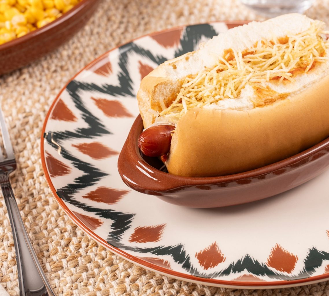 Imagem: Mesa posta com cachorro-quente servido em travessa marrom da linha Feijoada Oxford sobre prato Unni Boho, acompanhamentos ao fundo.