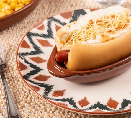Imagem: Mesa posta com cachorro-quente servido em travessa da linha Feijoada Oxford sobre prato Unni Boho, acompanhamentos ao fundo.