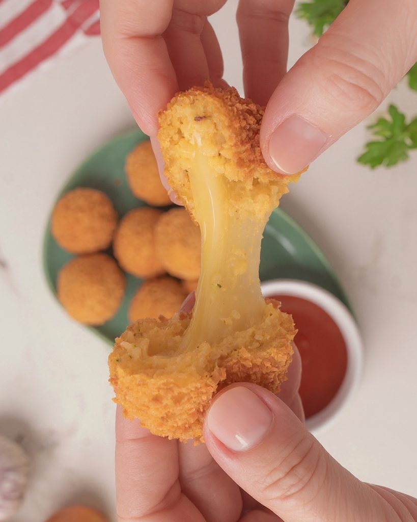 Close-up de bolinho de arroz recheado com queijo sendo aberto com as mãos, mostrando o queijo derretido puxando. Ao fundo, há mais bolinhos em um prato verde e um molho vermelho para acompanhar.