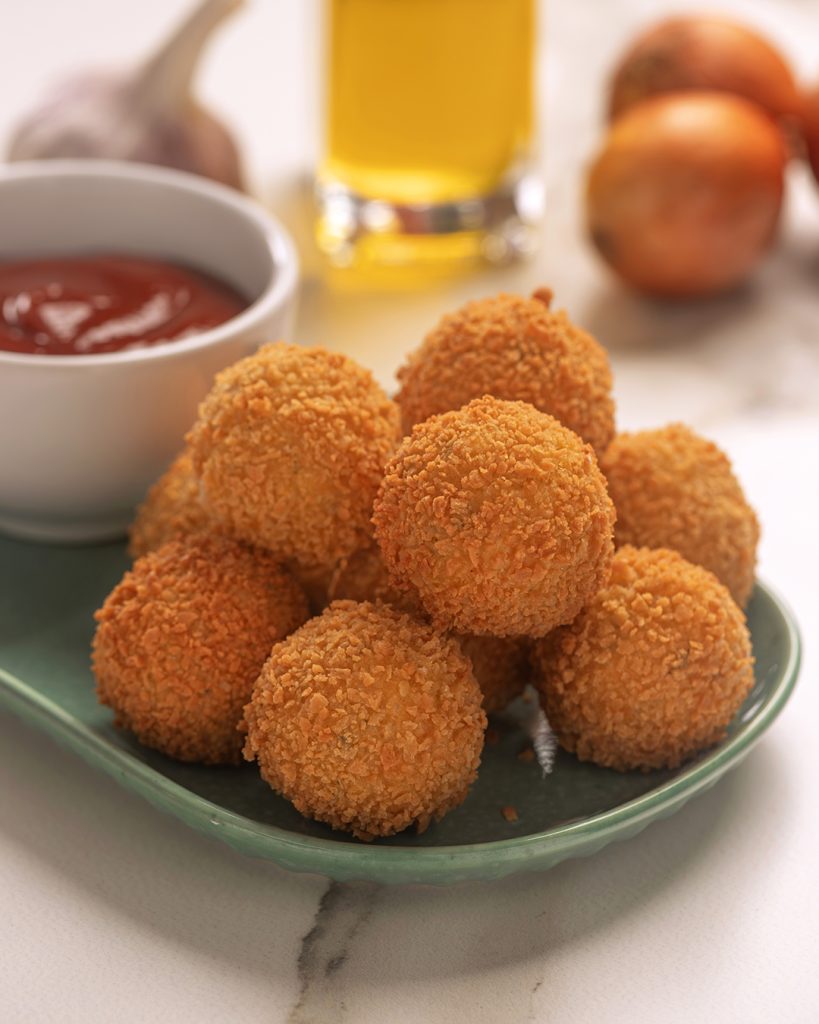 Bolinhos de arroz empanados e crocantes servidos em prato verde, acompanhados de molho vermelho. Ao fundo, ingredientes como alho, cebola e um recipiente com óleo desfocados.