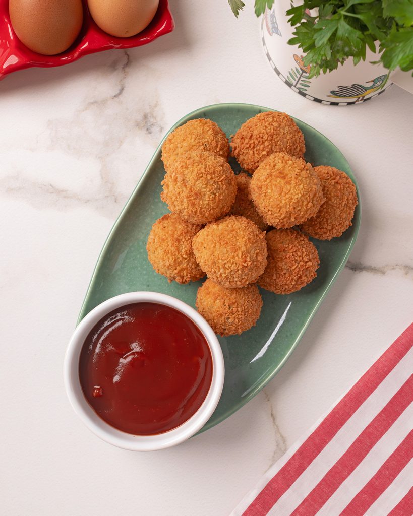 Prato verde com bolinhos de arroz empanados e crocantes acompanhados de molho vermelho. Ao fundo, elementos decorativos de cozinha sobre bancada clara.