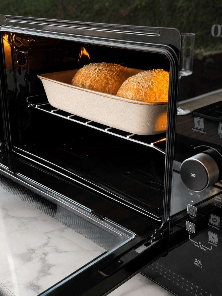 Cheeseburgers Wellington dourados e cobertos com gergelim assando no forno dentro de uma travessa refratária bege. A cena destaca a massa folhada inflada e crocante durante o preparo, com iluminação quente que valoriza o aspecto artesanal e apetitoso da receita.
