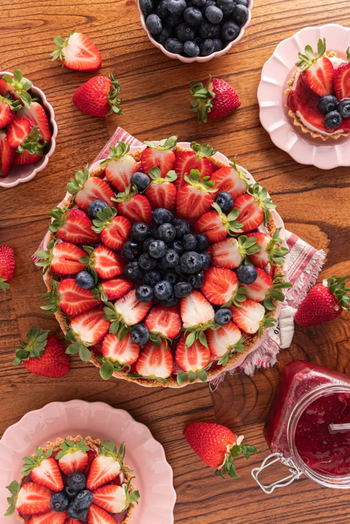 Torta decorada com morangos fatiados e mirtilos ao centro, servida sobre pratos na cor rosa claro, sobre mesa de madeira. Ao redor da torta há morangos inteiros, um pote com frutas vermelhas e um vidro de geleia, criando uma composição aconchegante e sofisticada.