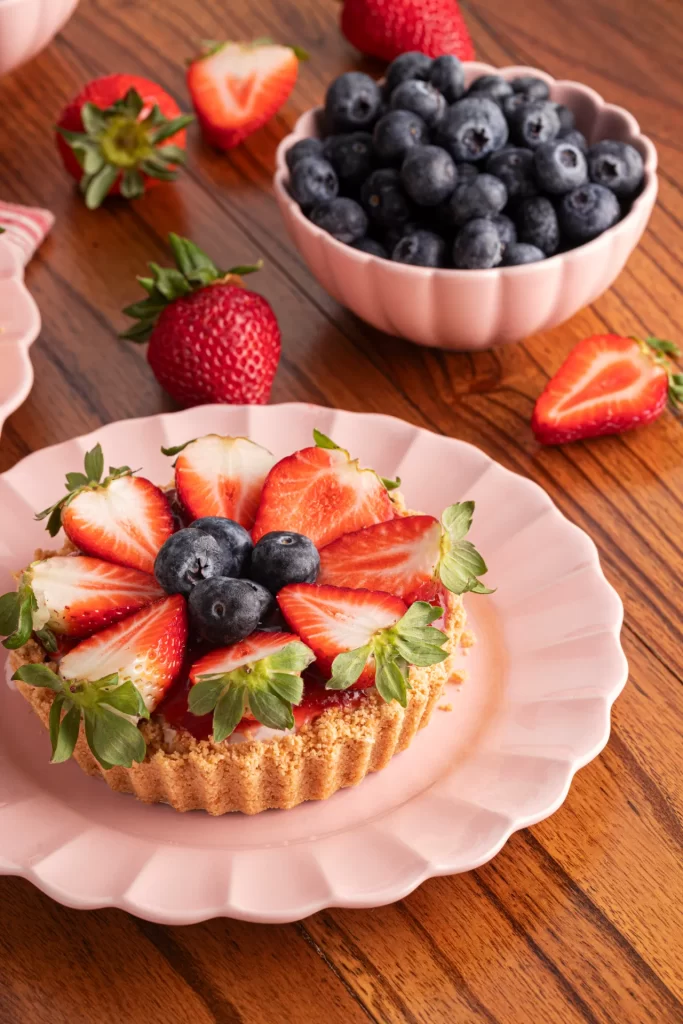 Mini torta de frutas vermelhas servida em prato rosa com bordas onduladas, decorada com morangos fatiados e mirtilos. Ao fundo, há uma tigela com mirtilos e morangos espalhados sobre a mesa de madeira.