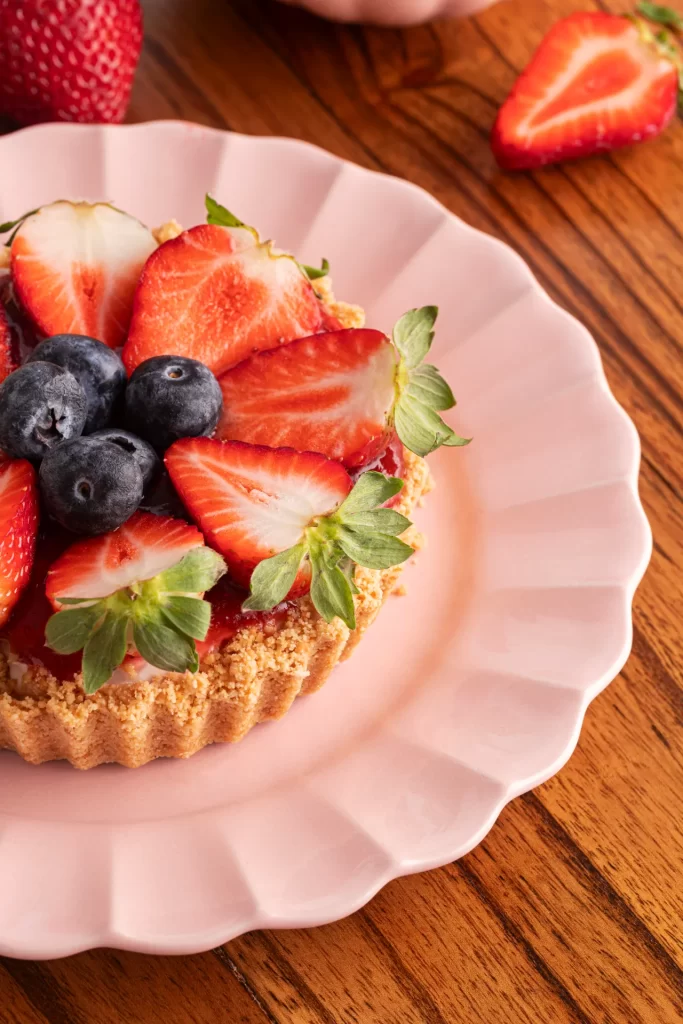 Close em mini torta decorada com morangos frescos e mirtilos sobre prato rosa delicado. A imagem destaca a textura da massa crocante, das frutas frescas e o fundo em madeira rústica.