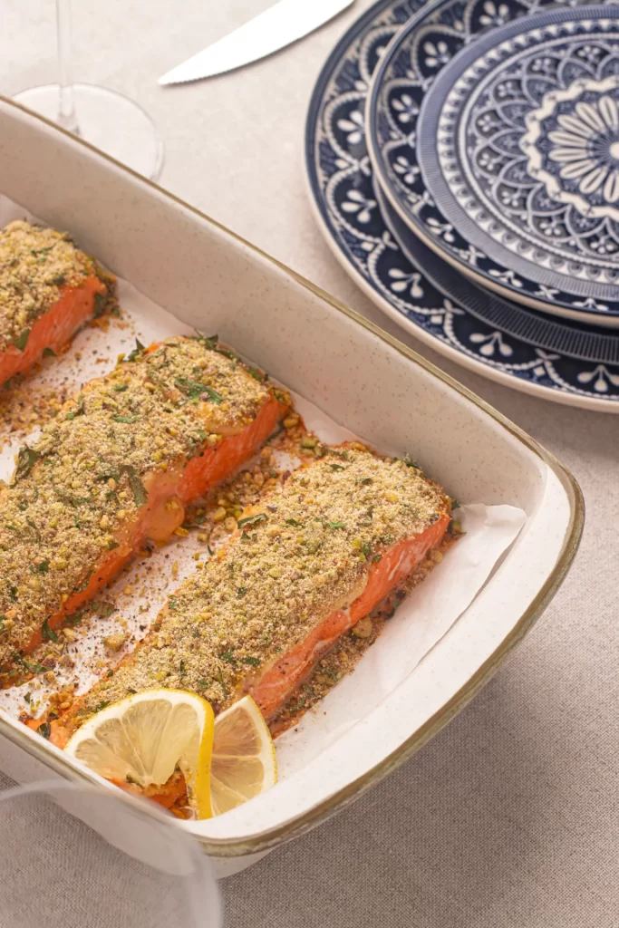 Travessa com pedaços de salmão assado com crosta crocante de pistache, decorados com fatias de limão, sobre uma mesa posta com pratos azuis estampados em estilo português.