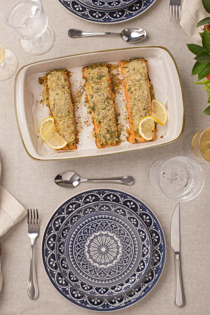 Mesa posta elegante com travessa de salmão com crosta de pistache ao centro, pratos azuis decorativos, talheres prateados e detalhes delicados em tons neutros.
