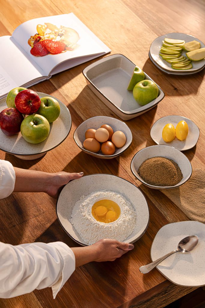 Pessoa preparando a massa da cuca em tigela sobre mesa de madeira, cercada por ingredientes como maçãs, ovos, manteiga, açúcar e farinha. Ao fundo, pratos com frutas, livro com figura de morangos aberto sobre a mesa e utensílios compõem um ambiente acolhedor de cozinha.