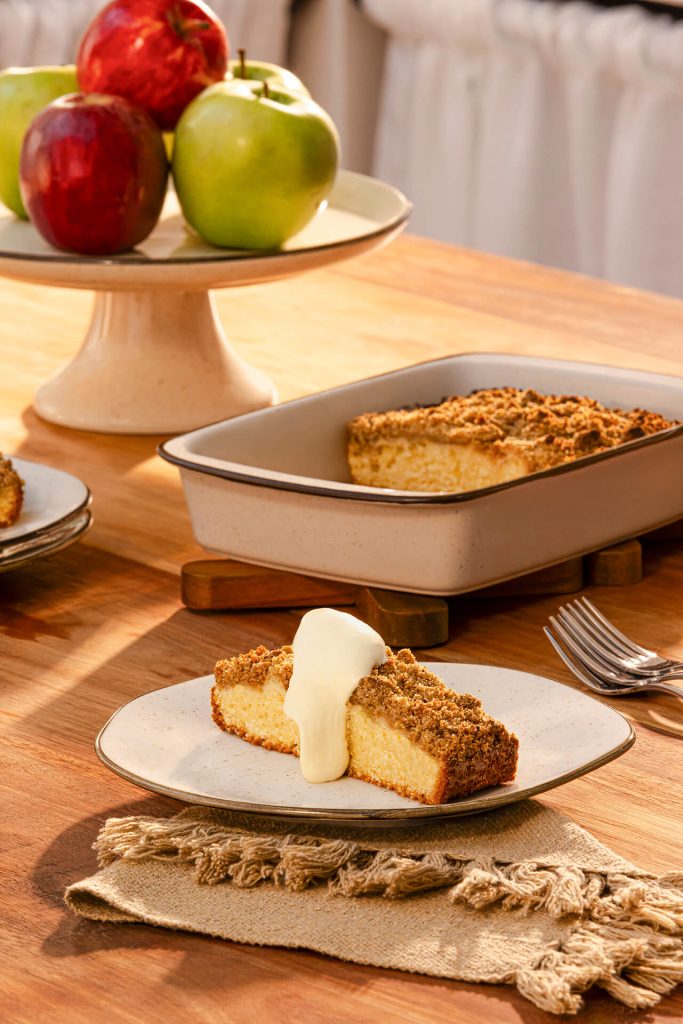 Fatia de cuca com massa fofinha e cobertura crocante servida em prato branco, acompanhada de creme claro ao lado. Ao fundo, forma com a cuca sobre mesa de madeira e fruteira com maçãs decorando o ambiente aconchegante.