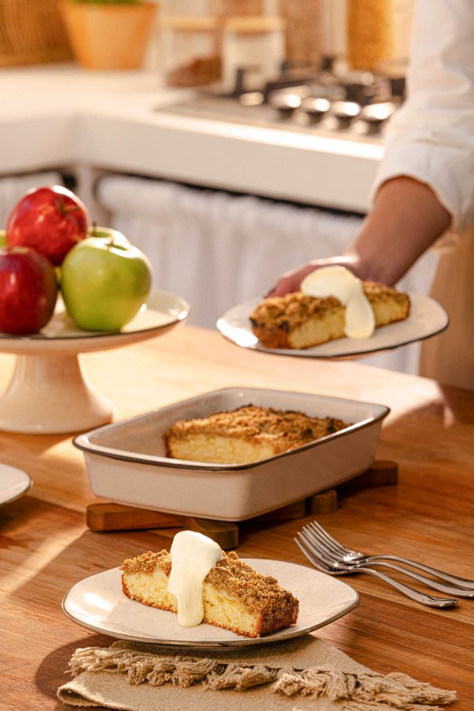 Fatia de cuca com farofa crocante servida com creme branco por cima, sobre uma mesa de madeira. Ao fundo, uma travessa com a cuca, maçãs decorativas e uma pessoa segurando um prato com outra fatia da sobremesa.