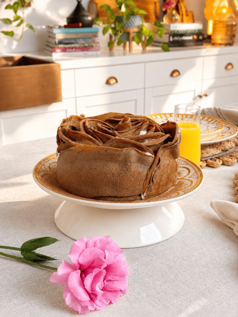 Bolo de rosa de panquecas de chocolate apresentado em uma boleira branca. Ao redor, elementos de café da manhã, como suco de laranja, guardanapo de tecido e flores, criam um ambiente aconchegante e iluminado.