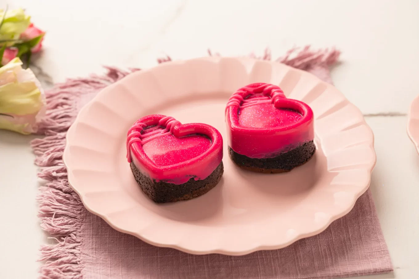Close-up focado nos dois mini brownies em formato de coração dispostos sobre o prato rosa ondulado. É possível ver o contraste entre a base escura do brownie de chocolate e a cobertura rosa brilhante no topo. Ao fundo, de forma desfocada, aparecem as flores e o jogo americano lilás.