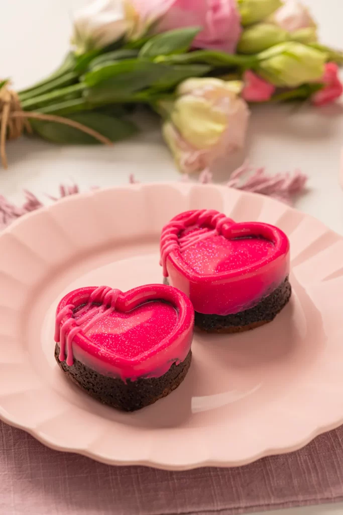 Mini brownies em formato de coração cobertos com uma camada rosa brilhante e decorados com fios de cobertura. Os doces estão servidos em um prato rosa claro, sobre uma superfície com tecido delicado. Ao fundo, um buquê de flores em tons de rosa e branco completa a composição romântica, criando um cenário perfeito para o Dia dos Namorados.