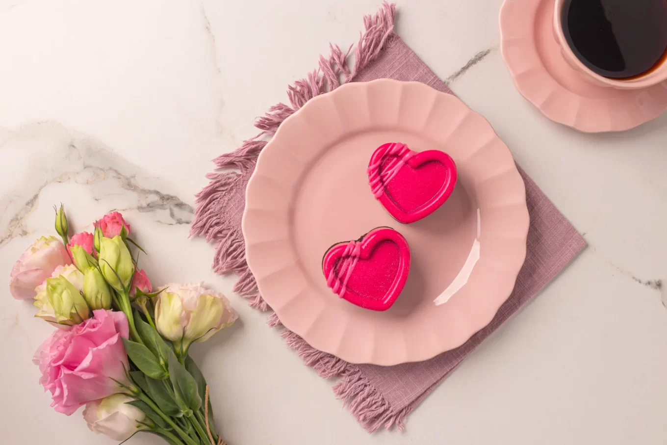 Foto em ângulo diagonal mostrando a mesma composição de café da manhã. O prato rosa com os dois mini brownies em formato de coração rosa choque está centralizado no jogo americano lilás. O arranjo de flores rosas e a xícara de café rosa aparecem nas laterais, complementando a decoração delicada.