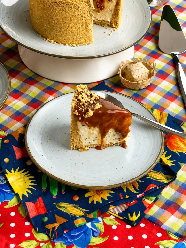 Fatia de torta de paçoca com doce de leite servida em prato de cerâmica clara. Ao fundo, a torta inteira aparece sobre uma boleira branca. A mesa está decorada com toalha xadrez colorida e tecido floral em tons de azul, amarelo e vermelho, criando um clima típico de festa junina.
