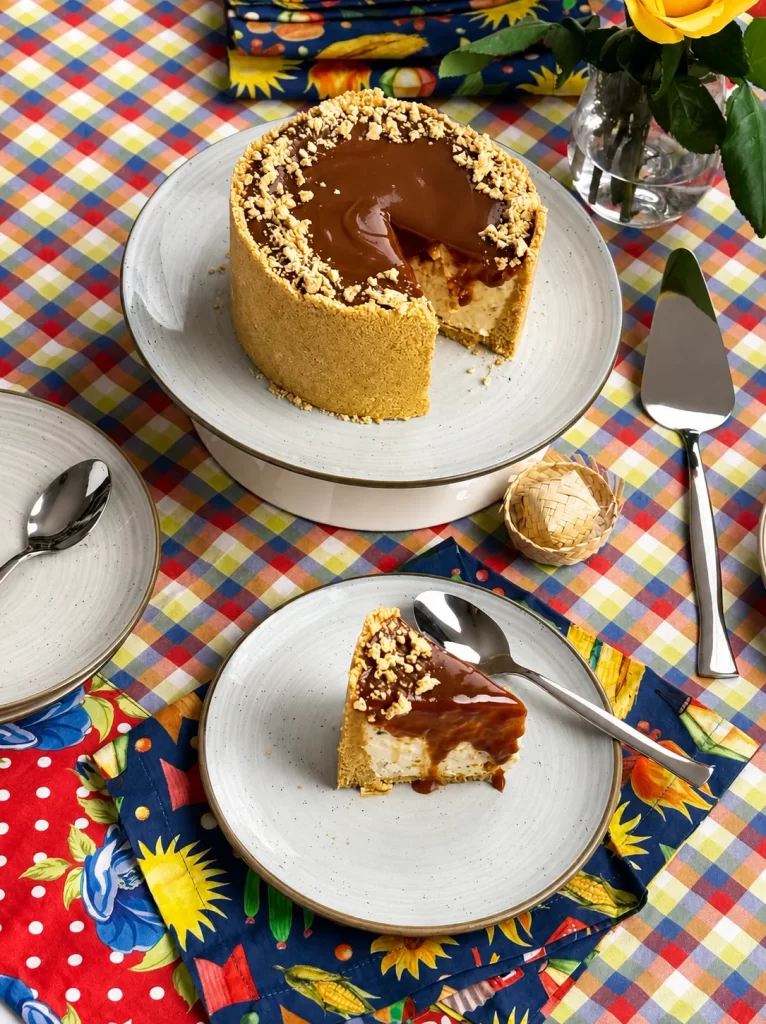 Torta de paçoca com recheio cremoso de doce de leite e uma fatia removida, revelando seu interior. Em primeiro plano, uma fatia é servida em prato de cerâmica claro. A composição inclui toalha xadrez colorida, tecido floral e elementos decorativos inspirados nas festas juninas.