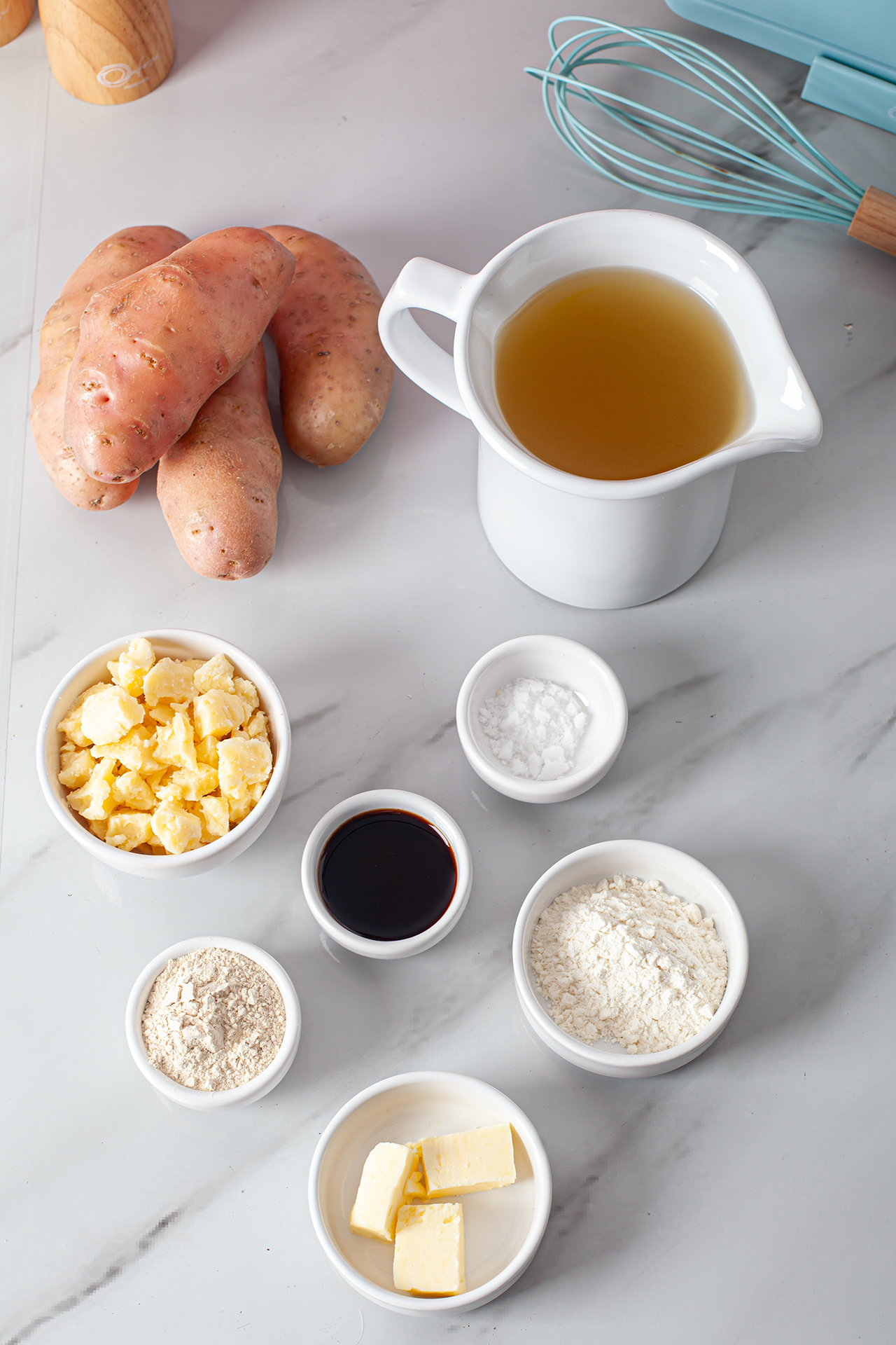 Ingredientes da receita de poutine organizados sobre uma bancada clara. Entre eles, estão batatas, caldo em uma jarra, queijo em cubos, molho escuro, farinha, manteiga e amido de milho em pequenos recipientes. Além disso, um fouet azul completa a composição.