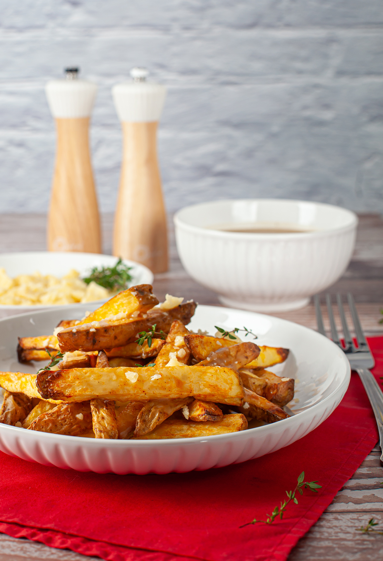 Prato de poutine com batatas assadas douradas cobertas por cubos de queijo e ervas frescas. Ao fundo, aparecem moedores de tempero, uma tigela de molho e talheres, enquanto o guardanapo vermelho adiciona um toque acolhedor à cena