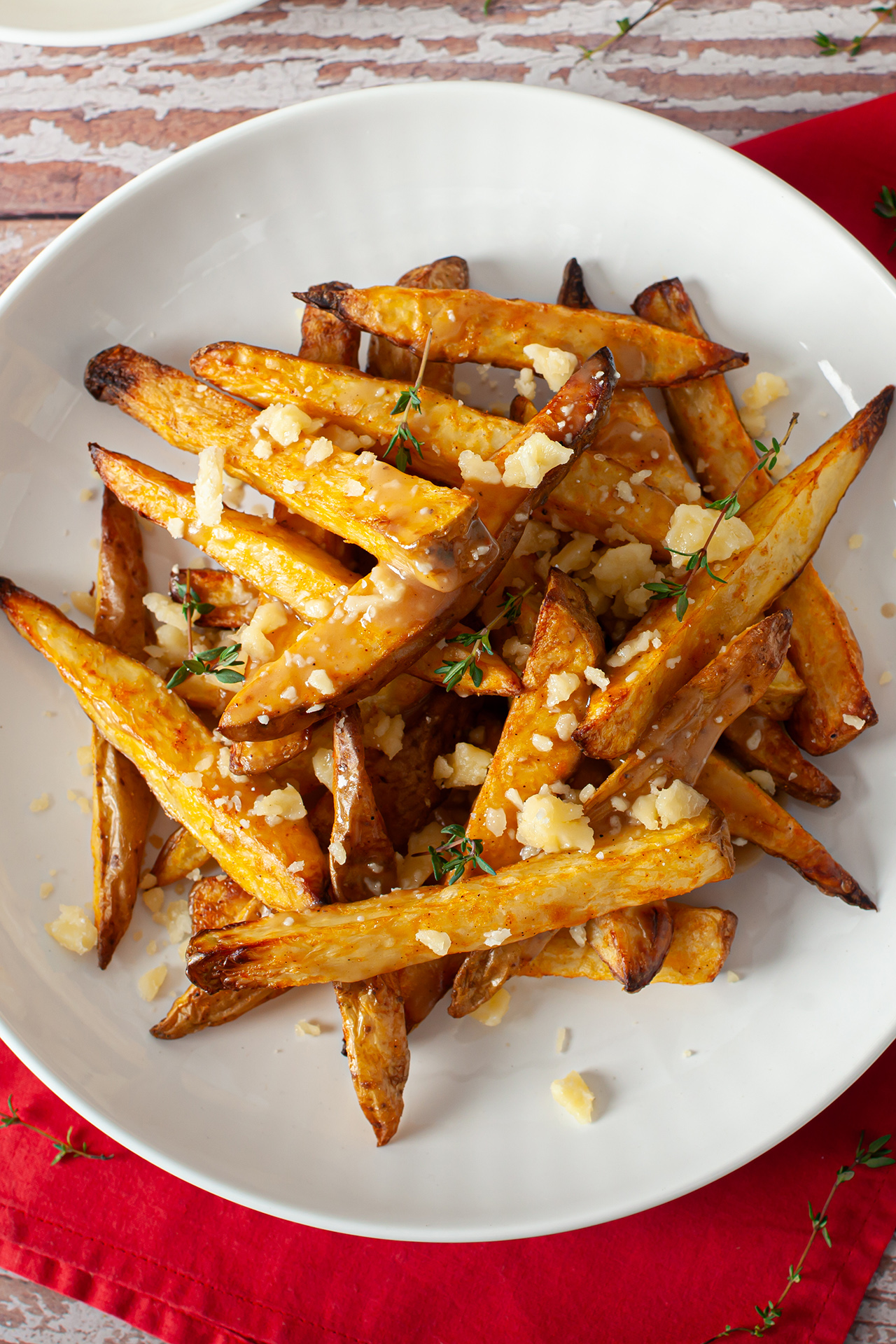 Vista superior de um prato de poutine com batatas rústicas assadas, cobertas por queijo em cubos levemente derretido e folhas de tomilho fresco. Enquanto o prato branco destaca os ingredientes, o guardanapo vermelho cria contraste com a mesa de madeira ao fundo.