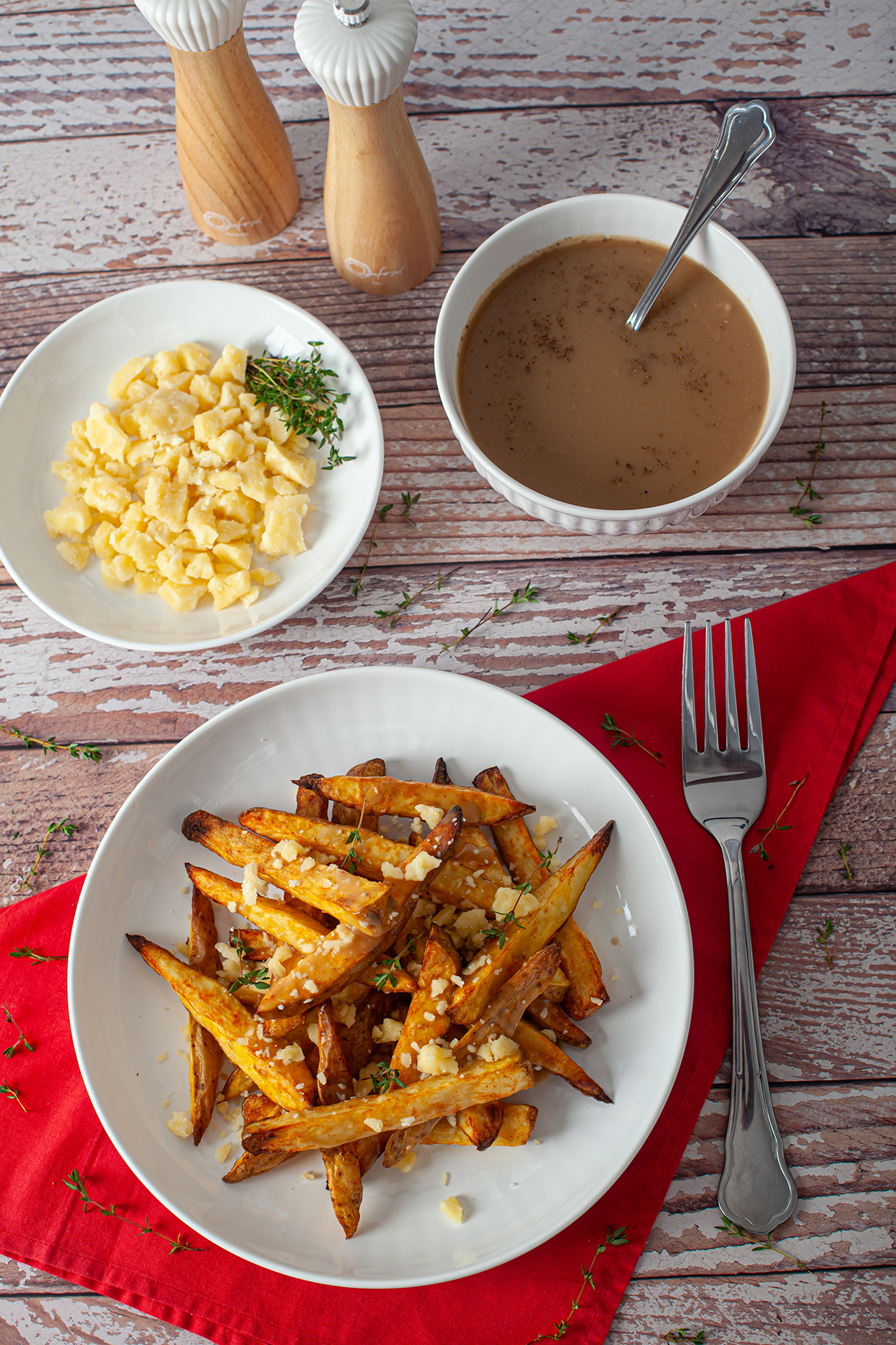 Prato de poutine servido sobre um guardanapo vermelho, com batatas rústicas douradas cobertas por queijo em cubos e molho. Ao fundo, além de uma tigela com molho marrom, há também um prato com queijo, tudo disposto sobre uma mesa de madeira rústica.