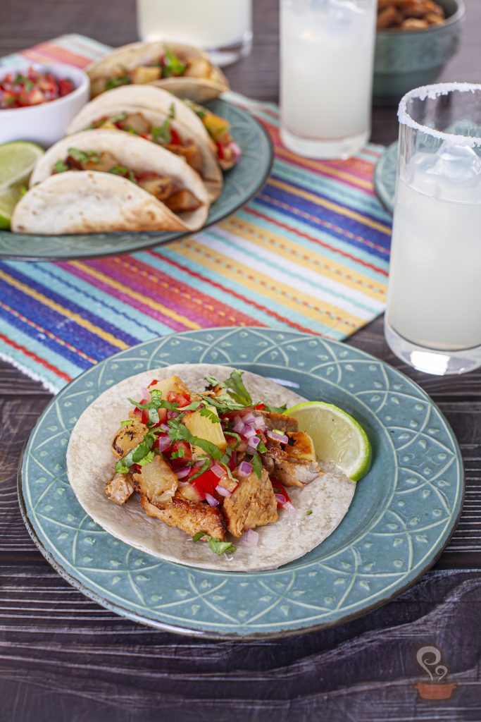 Close-up de um taco al pastor aberto sobre um prato de cerâmica azul. O recheio combina carne de porco grelhada, cubos de abacaxi caramelizado, tomate, cebola roxa e coentro. Ao fundo, aparecem outros tacos desfocados e copos com bebida cítrica gelada, criando uma atmosfera inspirada na culinária mexicana.