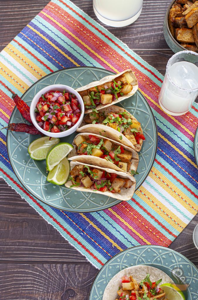 Vista superior de três tacos al pastor servidos em um prato de cerâmica azul. As tortilhas são recheadas com carne suína dourada, abacaxi grelhado, tomate, cebola roxa e coentro fresco. Ao lado, há uma tigela de pico de gallo, gomos de limão e pimentas secas. A composição está sobre uma toalha listrada colorida, com copos de bebida cítrica gelada ao redor.