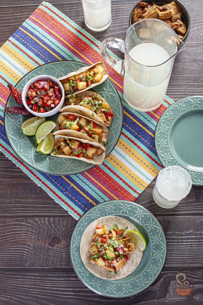 Vista aérea de uma mesa posta com tacos al pastor servidos em pratos de cerâmica azul. Os tacos levam carne de porco grelhada, abacaxi, tomate, cebola roxa e coentro. A composição inclui uma tigela de pico de gallo, gomos de limão, pimentas secas, uma jarra de bebida cítrica gelada e uma toalha listrada multicolorida que destaca o visual mexicano da receita.
