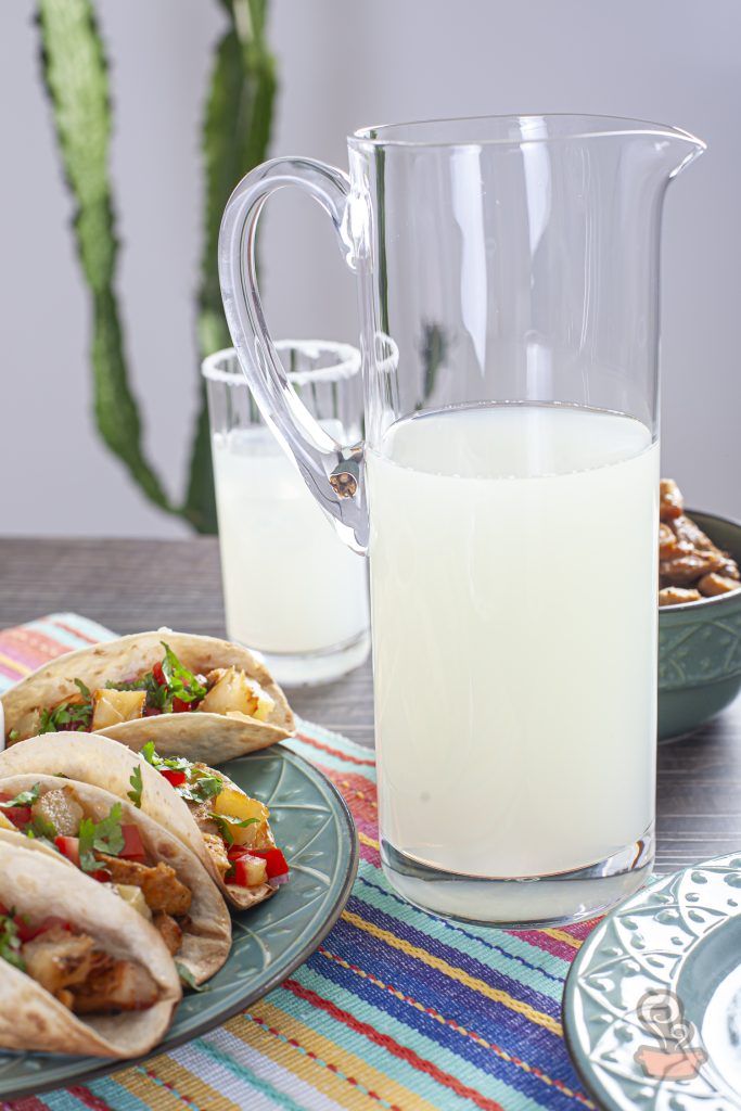 Jarra de bebida cítrica gelada em destaque sobre uma mesa de madeira escura, acompanhada por tacos al pastor servidos em pratos azuis. Os tacos são recheados com carne suína, abacaxi grelhado e salsa fresca. A toalha listrada colorida e os detalhes da mesa remetem ao clima vibrante da culinária mexicana.
