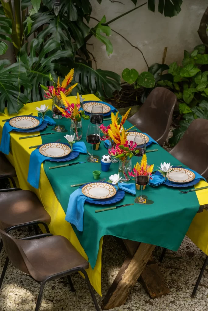Visão aérea mais ampla e aberta de toda a mesa retangular de madeira, montada em um ambiente externo cercado por plantas e folhagens. A mesa está vestida com uma sobreposição de toalhas amarela e verde, acomodando seis lugares completos com a louça decorativa azul e branca, guardanapos azuis e talheres coordenados. Cadeiras de design moderno em tom marrom escuro estão dispostas ao redor da mesa.