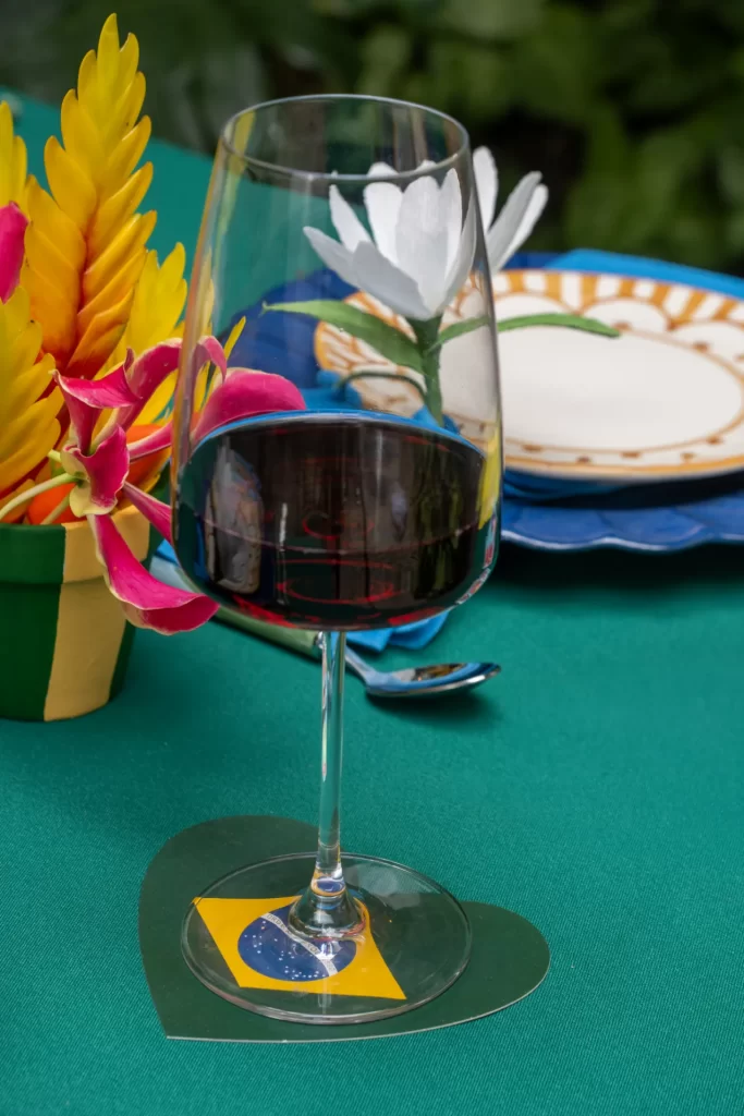 Uma taça de vinho tinto repousa sobre um descanso de copo verde em formato de coração, que traz a bandeira do Brasil estampada no centro. A taça está posicionada sobre uma mesa com toalha verde-bandeira, decorada ao fundo com flores tropicais amarelas e rosa, além de um prato de louça com detalhes em azul e amarelo.