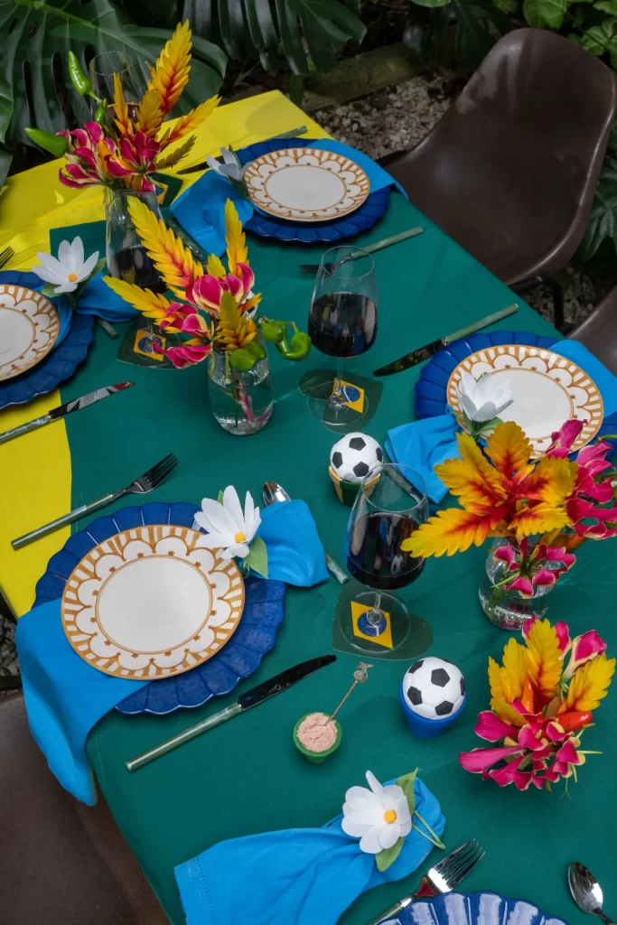 Ângulo diagonal que mostra parte da mesa decorada para duas ou mais pessoas. Os lugares seguem a mesma composição com pratos brancos e dourados, sousplats azuis e guardanapos com flores brancas. Vasos de vidro transparentes exibem arranjos de flores tropicais em tons de amarelo, vermelho e rosa. Pequenas réplicas de bolas de futebol decoram o centro da mesa verde e amarela.