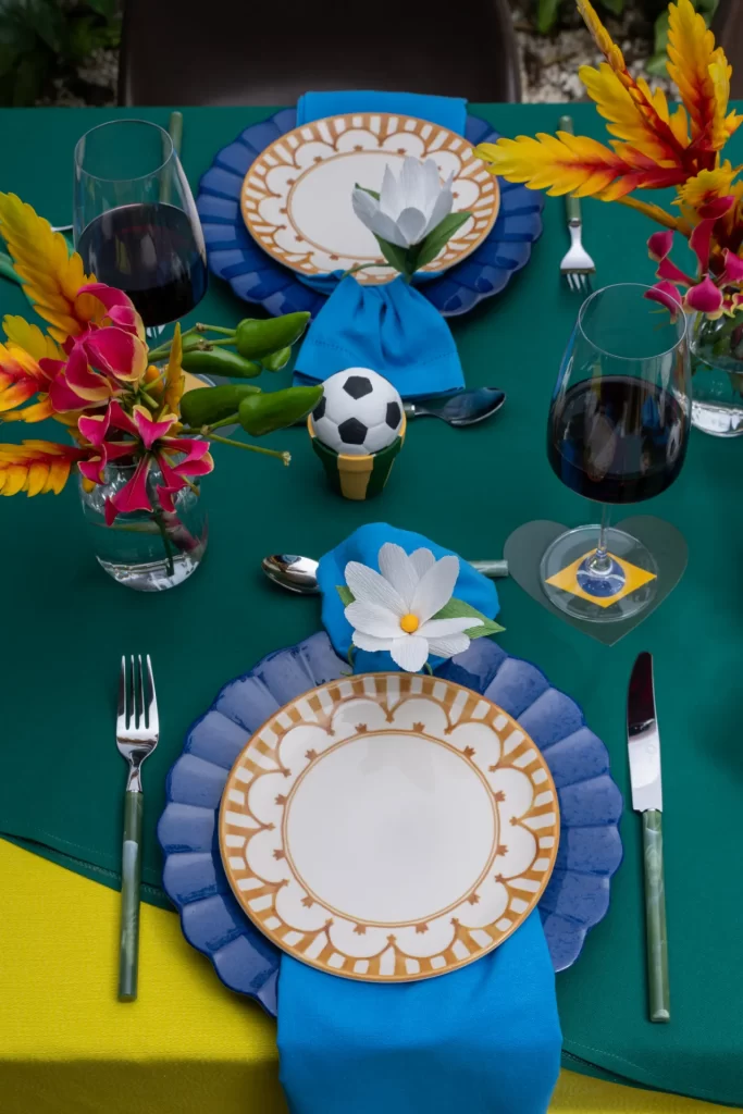 Vista aproximada de um lugar montado à mesa. Sobre uma toalha verde-bandeira com base amarela, destaca-se um prato de porcelana branca com bordas ornamentadas em dourado, posicionado sobre um sousplat azul com bordas onduladas. Um guardanapo de tecido azul vibrante está preso por um anel em formato de flor branca. Garfo e faca com cabos verdes ladeiam o prato. Próximo a eles, há uma mini bola de futebol decorativa e taças de vinho com descansos temáticos da bandeira do Brasil, além de arranjos com flores tropicais coloridas.