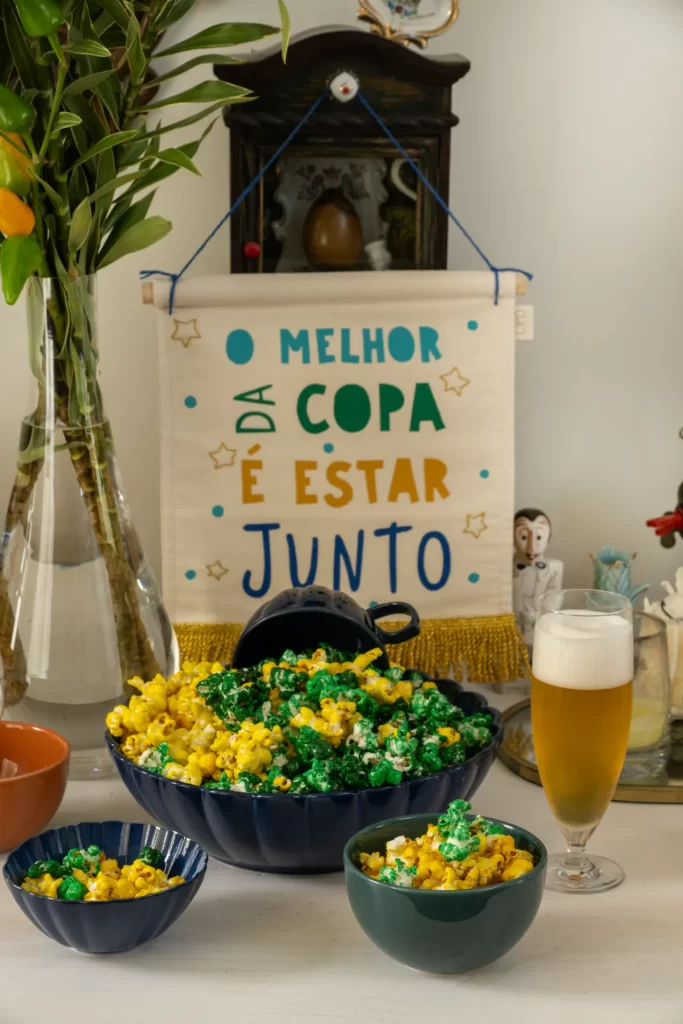 Uma mesa decorada traz em destaque uma grande tigela de louça azul-escura cheia de pipocas coloridas de verde e amarelo, com uma xícara azul virada no topo. À frente, há duas tigelas menores também servidas com a pipoca temática. Ao lado direito, destaca-se um copo alto de cerveja com colarinho de espuma branca. Ao fundo, uma flâmula de tecido branco pendurada exibe a frase decorativa escrita em letras coloridas: "O melhor da Copa é estar junto". À esquerda, um vaso de vidro alto com folhagens verdes complementa a ambientação.