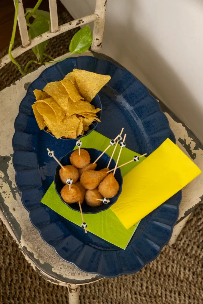 Travessa oval azul da coleção Flora Amor-Perfeito servindo mini coxinhas em palitos decorados com bolas de futebol e chips de milho. A composição conta com guardanapo verde-limão, formando uma proposta prática e descontraída para receber amigos durante os jogos da Copa do Mundo.