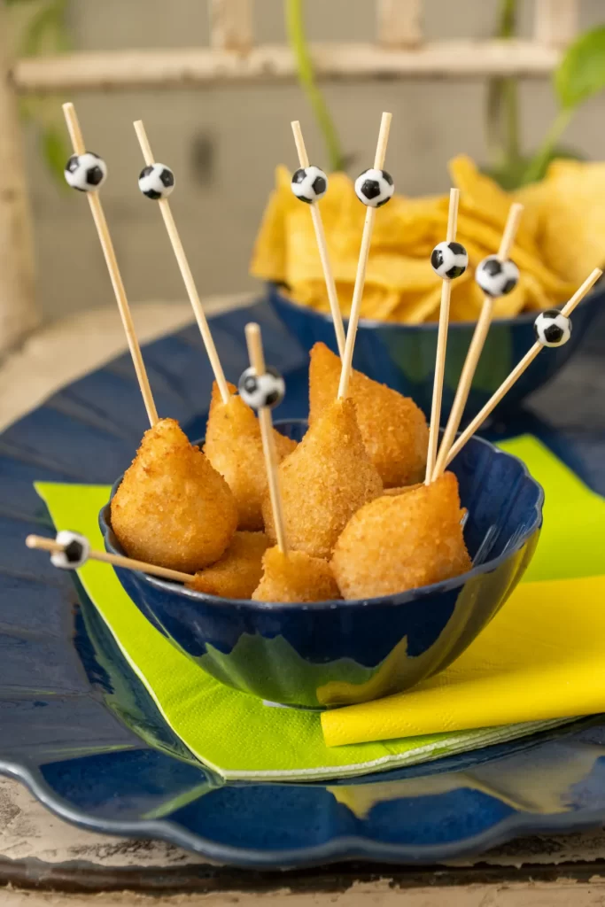 Tigela de sobremesa azul da coleção Flora Amor-Perfeito com mini coxinhas servidas em palitos decorados com bolas de futebol. A composição está sobre um prato azul da mesma coleção, acompanhado de guardanapo verde-limão, criando uma mesa temática para assistir aos jogos da Copa do Mundo.