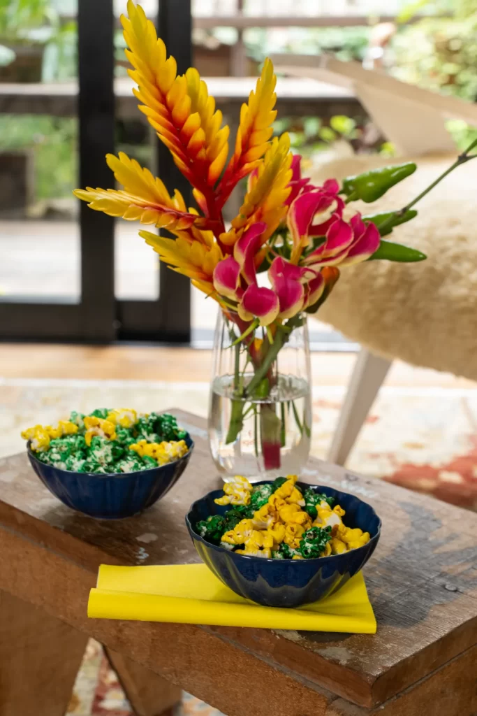 Um pequeno banco de madeira serve de apoio para duas tigelas azuis cheias de pipoca colorida artificialmente de verde e amarelo. Ao fundo, um vaso de vidro transparente exibe um arranjo alto com flores tropicais vermelhas, amarelas e folhagens.