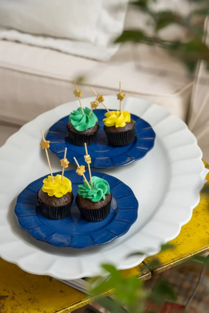 Uma travessa oval branca serve de apoio para dois pequenos pratos azuis, cada um contendo dois cupcakes de chocolate. Os bolinhos são cobertos com glacê nas cores amarela e verde, e decorados com palitos que trazem pequenas estrelas douradas no topo.