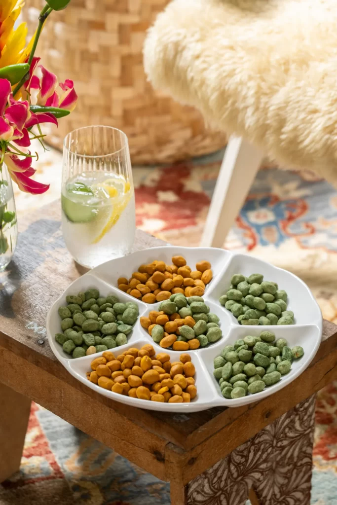 Um petisqueira redonda e branca com divisórias está posicionada sobre um banco rústico, servida com amendoins crocantes nas cores verde e amarela. Atrás do prato, há um copo alto com água aromatizada, rodelas de limão e pepino, além de um arranjo floral colorido.