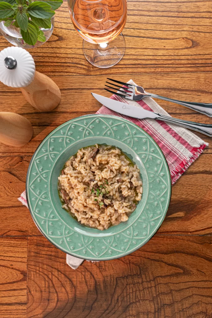 Risoto de file servido em um prato verde, com texturas nas bordas. O prato está em cima de um tecido vermelho e branco, em uma mesa de madeira. Na imagem, além do prato aparece um garfo, uma faca, saleiro, moedor de pimenta, uma taça de vinho branco e um pequeno vaso transparente com uma planta verde.