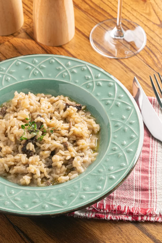 Foto do risoto de filé já pronto, servido em um prato na cor verde água, com texturas nas bordas! Na foto com zoom, aparece também um pouco do garfo e da faca sobre a mesa de madeira, o tecido em baixo do prato, um pedaço da taça de vinho e uma parte do saleiro e moedor de pimenta.